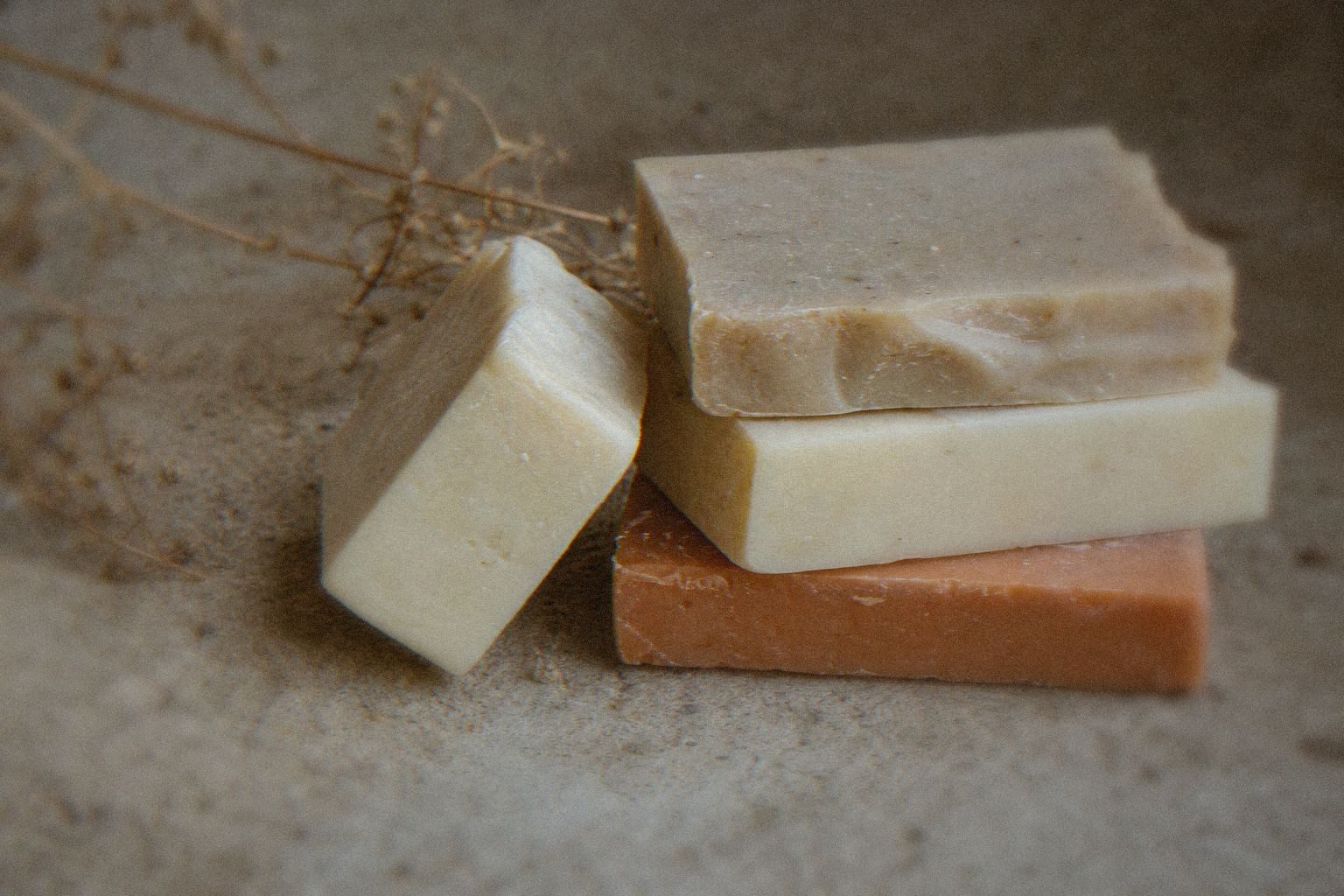 A stack of four handmade natural soap bars in cream, beige and terracotta tones, next to a sprig of dried flowers on a stone surface